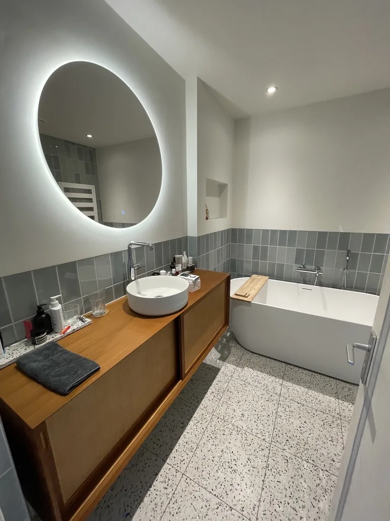 Dezelfde badkamer na de renovatie, met een terrazzovloer, een houten wastafelmeubel en een verlichte spiegel