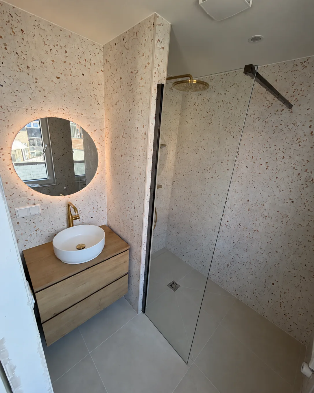 Badkamer met een terrazzowand, een ronde verlichte spiegel en een douche achter glas